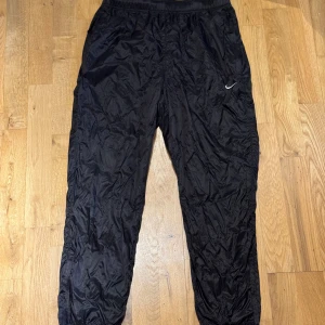 Svarta träningsbyxor från Nike - Snygga svarta träningsbyxor från Nike i 100% nylon. De har en elastisk midja och en liten vit Nike-logga på benet. Perfekta för träning eller en avslappnad dag. Strl M men passar mindre i storleken. 