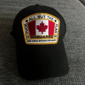 Svart keps från Dsquared2 - Snygg svart keps från Dsquared2 med ett broderat märke framtill som har ett rött lönnlöv och text. Justerbar rem baktill. Perfekt för en cool och avslappnad stil.