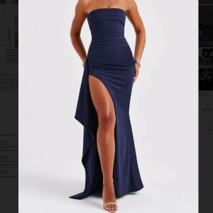 Mörkblå axelbandslös balklänning  - Elegant mörkblå axelbandslös klänning med en hög slits som ger en sofistikerad look. Perfekt för speciella tillfällen med sin figurnära passform och långa längd. Priset på klänningen var på hemsidan 1500 kr.