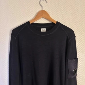 Mörkblå tröja från C.P. Company - Snygg mörkblå tröja från C.P. Company i merinoull. Tröjan har långa ärmar och en unik detalj med en lins på ärmen. Perfekt för en stilren och modern look. Använd cirka 3 gånger!