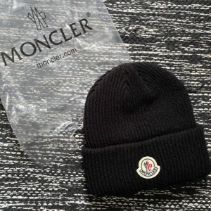 Svart Moncler mössa   - Snygg svart  mössa från Moncler med logo. Perfekt för kyliga dagar. Kommer i originalförpackning.