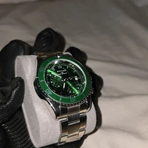 Grön klocka med silverarmband - Snygg klocka med grön urtavla och matchande grön bezel. Klockan har ett robust silverfärgat armband i metall. Perfekt för den som vill ha en iögonfallande accessoar. Datumet och tiden går att styra med kornan på klockan. Sido knappar är endast dekoration.