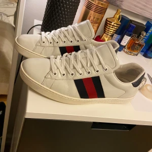 Gucci ace sneakers - Snygga vita sneakers från Gucci med klassiska ränder i rött och blått på sidan. Skorna har en stilren design med snörning och en diskret logga på hälen. Perfekta för en trendig look. JAG TAR BYTEN