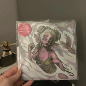 Milk of the Siren - Urban Outfitters exklusiv singel 'Milk of the Siren' av Melanie Martinez, utgiven 2024. Omslaget har en unik konstnärlig design med en fantasifigur i rosa och gröna toner. Perfekt för samlare och fans av Melanie Martinez.