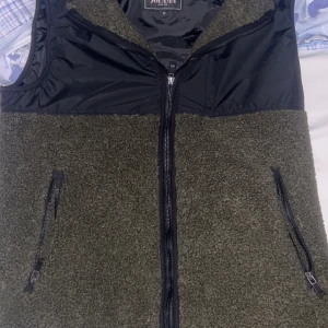 Olivgrön fleeceväst med dragkedja - Snygg olivgrön fleeceväst från Sofari med svarta detaljer. Västen har en dragkedja framtill och två praktiska fickor med dragkedjor. Perfekt för lager-på-lager under kyliga dagar.