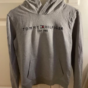 Grå hoodie från Tommy Hilfiger - Säljer en stilren grå hoodie från Tommy Hilfiger med klassisk logga på bröstet. Tröjan har en bekväm passform med luva. Perfekt för en avslappnad stil. Om ni har frågor skriv!