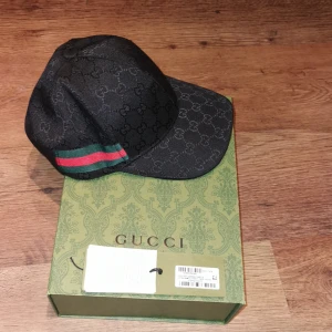 Svart keps från Gucci - Snygg svart keps från Gucci med det ikoniska GG-mönstret och röd-gröna ränder på sidan. Perfekt för att ge din outfit en lyxig touch. Kepsen kommer i originalförpackning.