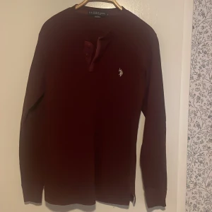 Vinröd tröja från Polo - Säljer en stilren vinröd tröja från Polo med knappar vid halsen och ett broderat logotypmärke på bröstet. Tröjan har långa ärmar och är perfekt för en avslappnad look. Kom DM kring några funderingar.