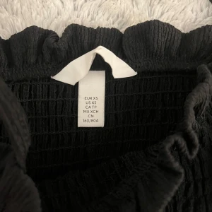 Svart långklänning - Elegant svart långklänning med smockdetaljer upptill. Perfekt för en stilren look. Klänningen har en vacker fall och är tillverkad i ett mjukt material som ger en bekväm passform.