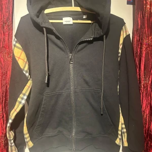 Svart burberry kofta  - Säljer en svart kofta från burberry med dragkedja och rutiga detaljer på ärmarna. Tröjan har en justerbar huva med snörning och praktiska fickor framtill. Perfekt för en avslappnad stil. Hör av er vid minsta fundering 🍾