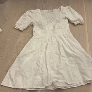 Vit klänning från NA-KD - Säljer en söt vit klänning från NA-KD designad av Nicole Mazzocato. Klänningen har korta puffärmar och en v-ringad design med knappar framtill. Den är dekorerad med spetsdetaljer som ger en romantisk touch. Perfekt för sommarens alla tillfällen! Använd ett par fåtal gånger