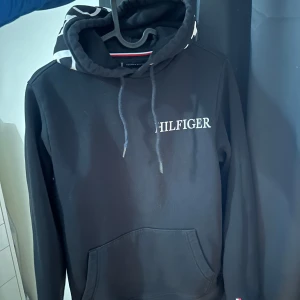 Svart hoodie från Tommy Hilfiger - Säljer en stilren svart hoodie från Tommy Hilfiger med texten 'HILFIGER' tryckt på bröstet och huvan.  Storlek XS