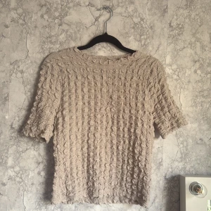 Beige tröja från Lindex - Säljer en beige tröja från Lindex med ett unikt texturerat mönster. Tröjan har korta ärmar och en rund halsringning. Perfekt för en stilren och bekväm look.