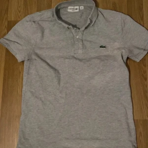 Grå pikétröja från Lacoste - Säljer en klassisk grå pikétröja från Lacoste med den ikoniska krokodilloggan på bröstet. Tröjan har korta ärmar och en knappslå framtill. Perfekt för en stilren och avslappnad look.