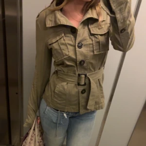 Militärgrön jacka med bälte - Snygg militärgrön jacka med knappar och bälte i midjan. Jackan har långa ärmar och flera fickor framtill. Perfekt för en stilren look.