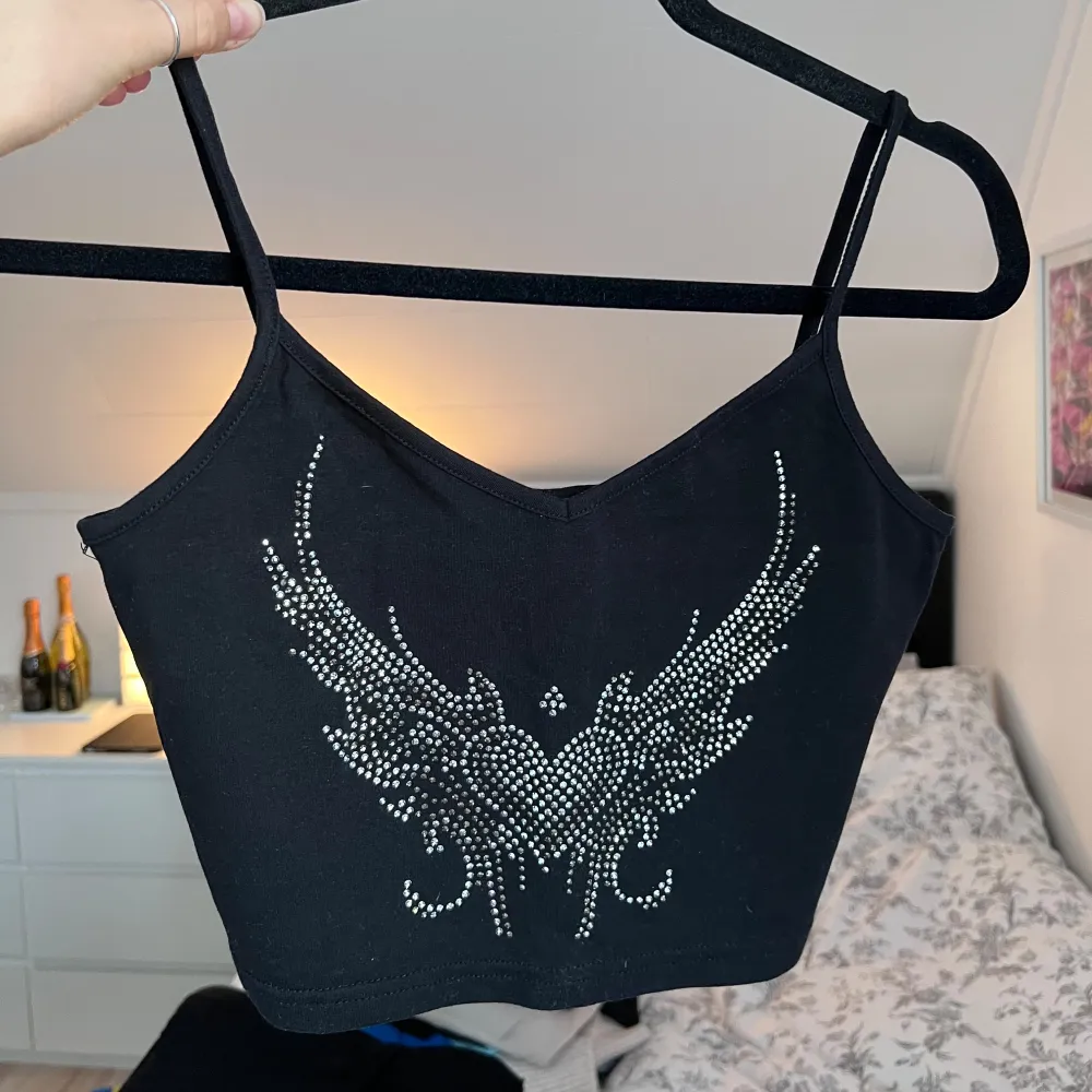 Snygg svart linnetopp med smala axelband och ett coolt vingmönster i silverfärgade stenar på framsidan. Perfekt för en trendig look. Aldrig använd. Önskar du veta exakta mått? Tveka inte att höra av dig!. Yläosat.