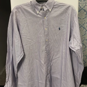 Randig skjorta från Ralph Lauren - Vi säljer nu en klassisk randig skjorta från Ralph Lauren i ljuslilla och vitt. Skjortan är långärmad med knappar och har en broderad logotyp på bröstet. Perfekt för en stilren look!