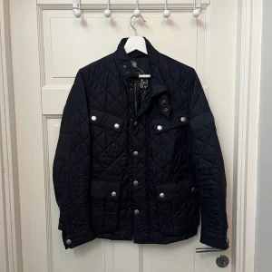 Svart quiltad jacka från Barbour - Säljer en stilren svart quiltad jacka från Barbour. Jackan har en hög krage med knappar och dragkedja framtill. Den har flera fickor med knappar. Perfekt för lite kyligare dagar.