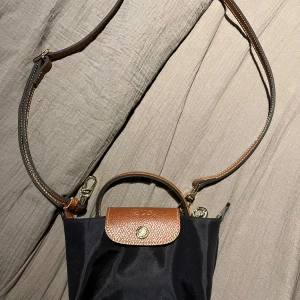 Svart och brun axelväska - Snygg svart axelväska med bruna detaljer och justerbar axelrem. Defekterna är att ringen på vänster sida är borta och att texten på öglorna är repade då man gjort bandbyten men inget som stör väskans funktion