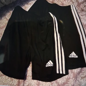 Svarta shorts från Adidas - Säljer ett par svarta Adidas shorts med de klassiska tre vita ränderna längs sidorna. Shortsen har elastisk midja och är tillverkade i ett lätt och bekvämt material, perfekt för sport och träning. Adidas-loggan är tryckt på framsidan.
