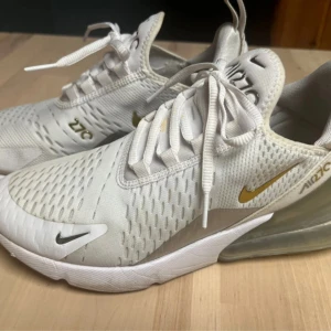 Nike Air Max 270 i vitt och guld - Snygga Nike Air Max 270 sneakers i vitt med guldiga detaljer. Skorna har en luftig design med snörning och en synlig Air Max-enhet i hälen för extra komfort. Perfekta för en sportig look.storlek 40.