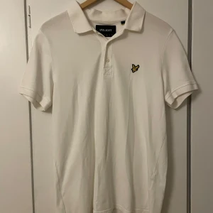 Vit pikétröja från Lyle & Scott - Säljer en klassisk vit pikétröja från Lyle & Scott med korta ärmar och en broderad gul logotyp på bröstet. Perfekt för en stilren och avslappnad look. Tröjan har en krage och knappar framtill.