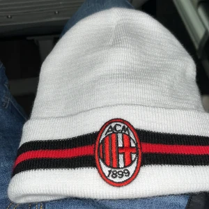 Vit mössa med AC Milan-logga - Snygg vit mössa med AC Milan-logga framtill. Mössan har svarta och röda ränder som ger en sportig look. Perfekt för fotbollsfans som vill visa sitt stöd. Pris kan diskuteras 