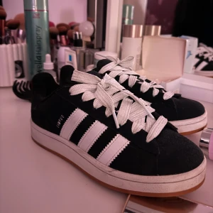 Svarta Adidas campus  - Snygga svarta Adidas sneakers med de klassiska tre vita ränderna på sidan. Skorna har vita snören och en vit sula med brun undersida. Perfekta för en avslappnad stil. Man får även med svarta skosnören 💞