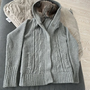 Grå stickad hoodie med päls luva - Mysig grå stickad hoodie med päls i luvan. Tröjan har ett flätmönster framtill och ribbade muddar vid ärmslut och nederkant. Perfekt för kyliga dagar nu till våren! Inga defekter på den och priset kan sänkas rejält vid snabb affär