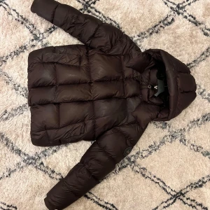 Brun dunjacka från Monel - Säljer en brun dunjacka från Monel med huva och dragkedja. Jackan har en quiltad design och är perfekt för kyligare väder. Märkeslogga på ärmen ger en extra touch. Passar bra för både stad och natur.