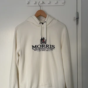 Vit hoodie från Morris i storlek  - Säljer en stilren vit hoodie från Morris med tryck 'Morris Sporting Club' och ett emblem på bröstet. Hoodien har en justerbar dragsko i huvan och långa ärmar. Perfekt för en avslappnad stil.