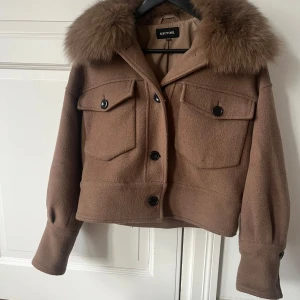 Brun jacka med pälsdetalj från Meotine  - Snygg brun jacka från Modström med en lyxig pälsdetalj på kragen. Jackan har knappar framtill och två stora fickor med lock. Perfekt för att hålla stilen i kyligare väder.