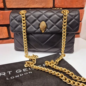 Svart quiltad axelväska från Kurt Geiger - Snygg svart axelväska från Kurt Geiger med quiltad design och guldfärgad kedja. Väskan har en unik detalj på framsidan och är perfekt för att ge din outfit en lyxig touch. Den är tillverkad i skinn och har en elegant och tidlös stil.