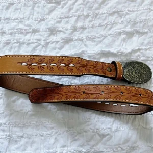 Brunt bälte med mönstrat spänne - Snyggt brunt bälte med ett detaljerat mönster och ett stort, ovalt spänne i metall. Perfekt för att ge en unik touch till din outfit. Aldrig använd så i nyskick. Storlek S. Full längd 74.5 cm (utan bältesspänne). Använd gärna KÖP NU om du vill köpa.