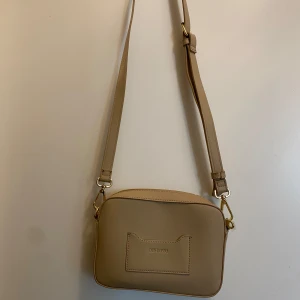 Beige axelväska från Don Donna - Snygg beige axelväska från Don Donna med justerbar axelrem och praktisk dragkedja. Väskan har en stilren design med en ficka på framsidan och guldfärgade detaljer. Perfekt för att bära dina nödvändigheter med stil. Den är i helt ny skick aldrig använd från Glitter! 