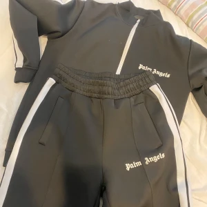 Svarta mjukisbyxor från Palm Angels - Snygga svarta mjukisbyxor från Palm Angels med vita sidoremsor och elastisk midja. Byxorna har en avslappnad passform och är perfekta för en casual look. Märkesloggan är tryckt på framsidan. Köpt på plick och kan inte försäkra äkthet därav priset. Scan finns men går inte alltid att scanna. Hör av för fler frågor och bilder