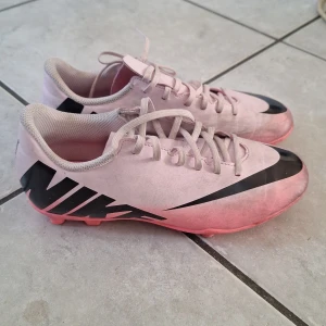 Rosa fotbollsskor från Nike - Säljer ett par rosa fotbollsskor från Nike med svarta detaljer och snörning. Skorna har en sportig design med dobbar för bra grepp på planen. Perfekta för fotbollsträning eller match.💗 ganska så använda som man kan se men inte slitna dobbar. Jag säljer de för att de är för små för mig.