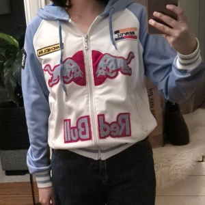 Vit och blå Red Bull jacka med luva - Säljer en cool vit och blå Red Bull jacka med luva och dragkedja. Jackan har flera patchar och logotyper, inklusive Red Bull och racinginspirerade märken. Perfekt för dig som gillar motorsport! 💙