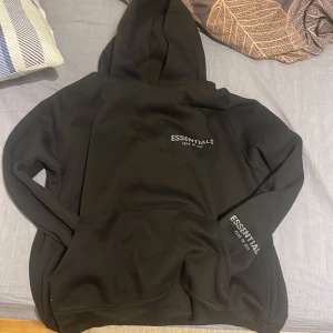 Svart hoodie från Essentials - Säljer en stilren svart hoodie från Essentials, Fear of God. Den har en klassisk design med en stor huva och en känguruficka framtill. Perfekt för en avslappnad look. Har krympt lite passar som S