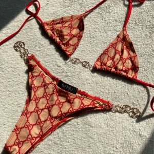 Beige och röd bikini med kedjedetaljer - Snygg beige bikini med röda detaljer och ikoniskt mönster. Både överdel och underdel har kedjedetaljer som ger en lyxig touch. Perfekt för sommarens alla badäventyr! Knappt använd då underdelen blivit för liten. Storlek M