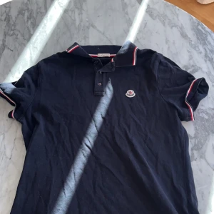  skorta Moncler  - Snygg svart pikétröja från Moncler med korta ärmar och klassisk krage. Tröjan har röda och vita detaljer på kragen och ärmsluten samt Monclers logga på bröstet. Perfekt för en stilren look.