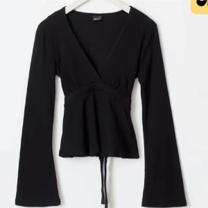 Svart omlottopp med långa ärmar - Snygg svart omlottopp med långa ärmar och v-ringning. Toppen har en knytfunktion i midjan för en justerbar passform. Perfekt för en stilren look. Endast provad, aldrig använd.