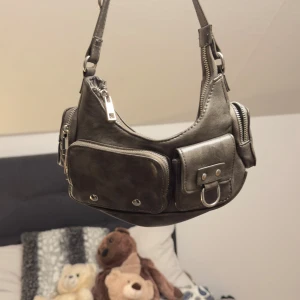 Vintage väska från Bershka - Snygg grå axelväska i skinn med flera praktiska fack och dragkedjor. Väskan har en justerbar axelrem och dekorativa metalldetaljer. Perfekt för att hålla ordning på dina saker med stil.