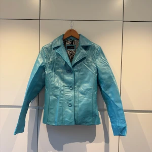 Turkos skinnjacka - Snygg turkos skinnjacka från Saki PULS med knappar framtill. Jackan har en klassisk krage och ett coolt svartvitt mönstrat foder. Perfekt för att ge din outfit en färgklick! Jackan är knappt använd och har inga defekter. Passar perfekt till våren som kommit tidigt i år! Pris kan diskuteras 