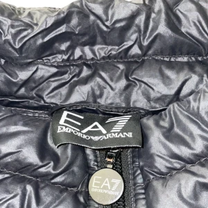 Svart väst från Emporio Armani - Snygg svart väst från Emporio Armani med dragkedja framtill. Västen har en quiltad design och är ärmlös, perfekt för lager-på-lager-stil. EA7-logga på bröstet ger en stilren touch.