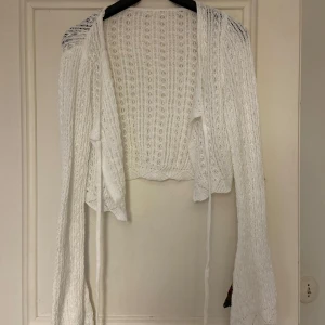 Vit stickad kofta med knytband - Säljer en vit stickad kofta med långa ärmar och knytband framtill. Koftan har ett fint mönster och är perfekt för lager-på-lager-stil. Den är både stilren och bekväm.