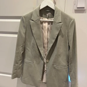 Grön kavaj från H&M - Snygg grön kavaj från H&M med klassisk design och långa ärmar. Kavajen har en enkel knapp framtill och två fickor med lock. Perfekt för en stilren look.