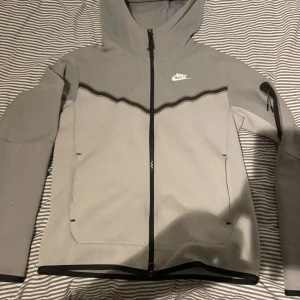 Grå jacka från Nike - Säljer en stilren grå jacka från Nike med dragkedja och huva. Jackan har en svart vågig detalj och är helt ny. den kostar egentligen 1500 kr men mitt pris är bara 600 kr 