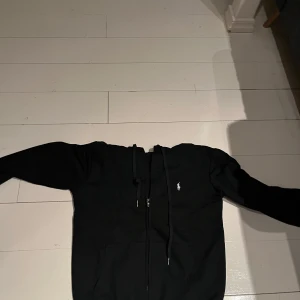 Svart hoodie från Ralph Lauren - Säljer en stilren svart hoodie från Ralph Lauren med dragkedja och det klassiska logotypbroderiet på bröstet. Perfekt för en avslappnad look. Den har långa ärmar och en praktisk huva med dragsko.