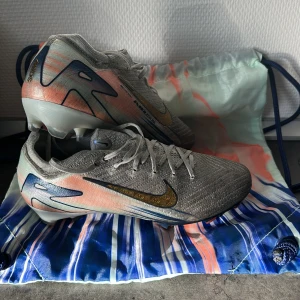 Nike fotbollsskor med grå och orange design - Snygga Nike Elit fotbollsskor i grått med orange och blå detaljer. Skorna har en modern design med snörning och en slitstark sula. Perfekta för fotbollsplanen med en stilren look.